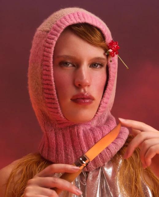 Close-up portrait of a woman wearing a pink knit balaclava holding a knife against a red-toned background. 