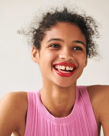 Portrait of a smiling woman in her 20s wearing a pink tank top with bright red lipstick against a plain white background