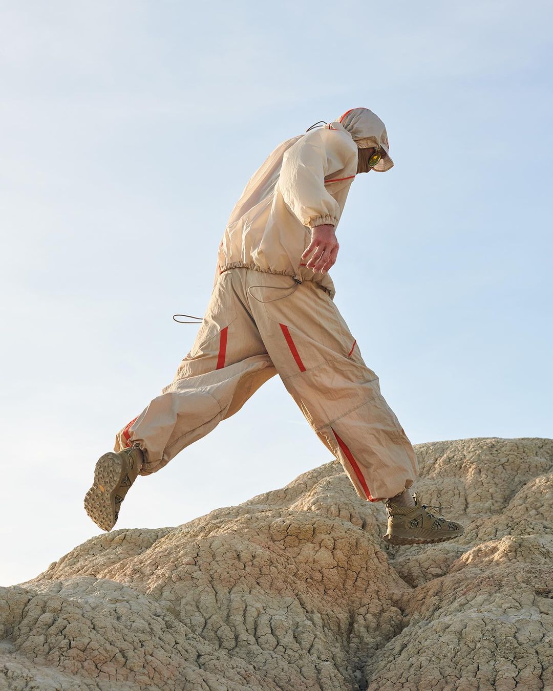Adult male climbs a textured beige hill wearing tan outdoor jacket, pants, and boots against pale blue sky in a medium outdoor shot.