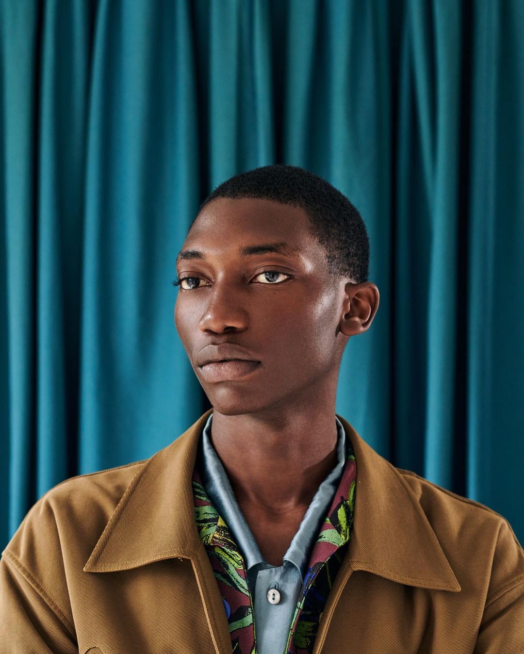 Portrait of a Black man with a serious expression in front of a turquoise draped fabric backdrop, wearing a brown jacket and patterned shirt.