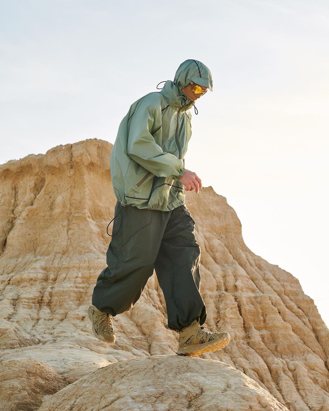A person in a hooded windbreaker and hiking boots walks in a desert landscape with a hill in the background against a bright sky.