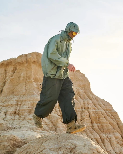 A person in a hooded windbreaker and hiking boots walks in a desert landscape with a hill in the background against a bright sky.