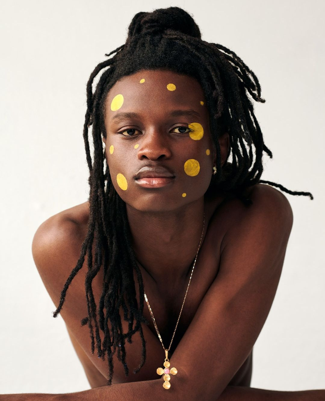 Close-up portrait of a Black male model with dreadlocks, yellow face paint, a gold cross necklace, shot in a photo studio on a white backdrop.