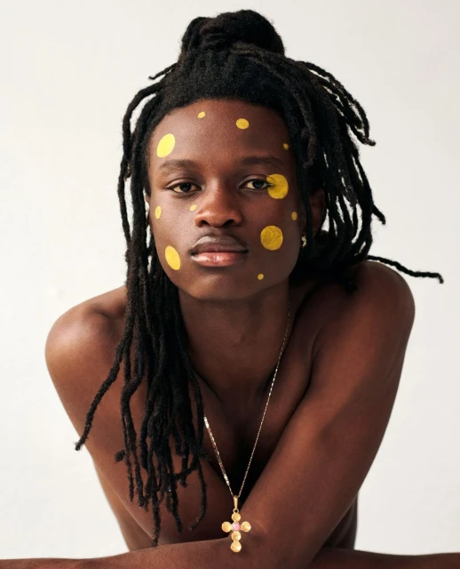 Close-up portrait of a Black male model with dreadlocks, yellow face paint, a gold cross necklace, shot in a photo studio on a white backdrop.