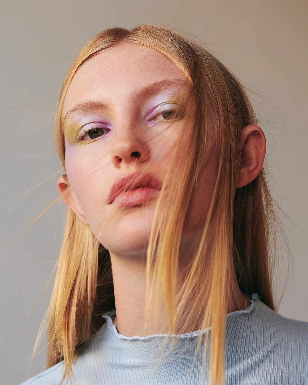 Close-up portrait of a blonde adult female with light skin, light blue top, light makeup with hues of lavender and yellow.
