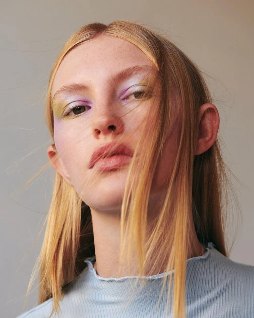 Close-up portrait of a blonde adult female with light skin, light blue top, light makeup with hues of lavender and yellow.