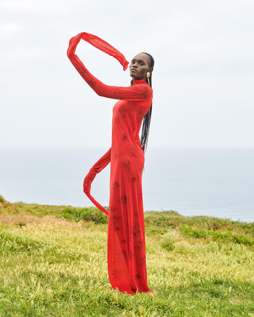 Fashion editorial shot of a Black woman in a long red patterned dress, on a grassy hill overlooking the sea, holding fabric in the air.