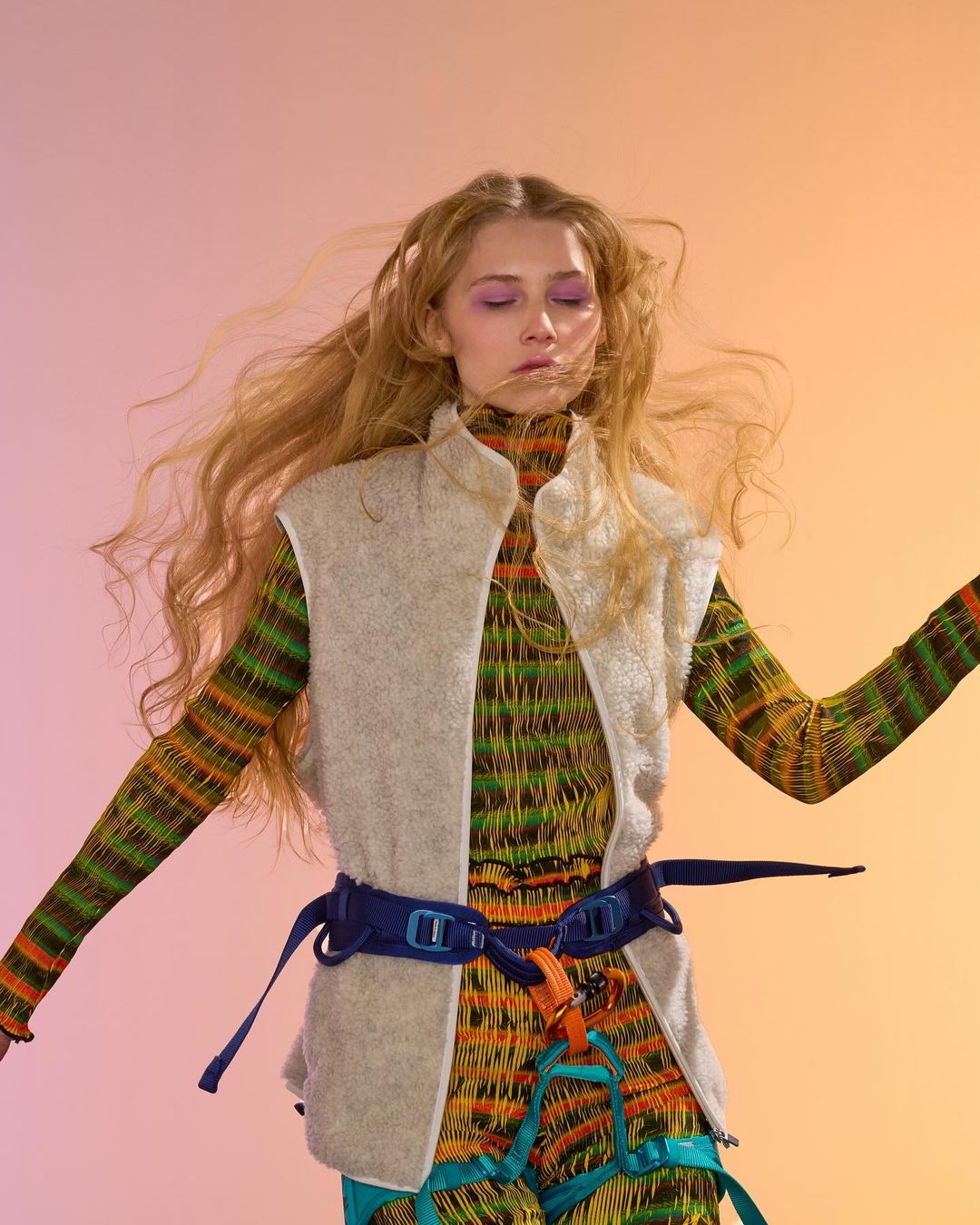 Young woman with long blond hair poses in an orange and pink gradient environment. She wears a vest and patterned jumpsuit.