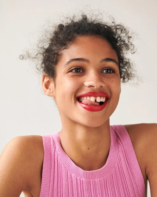 Close-up portrait of a smiling young adult female with curly hair and bright red lipstick sticking her tongue out slightly in a photo studio.