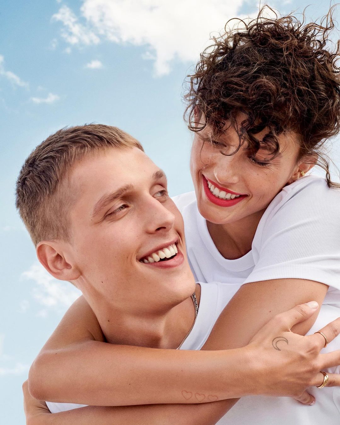 A young adult couple smiles together against a blue sky with clouds. The woman is on the man's back, hugging him in a happy embrace.