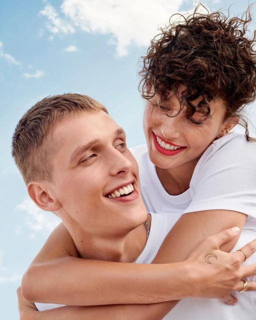 A young adult couple smiles together against a blue sky with clouds. The woman is on the man's back, hugging him in a happy embrace.