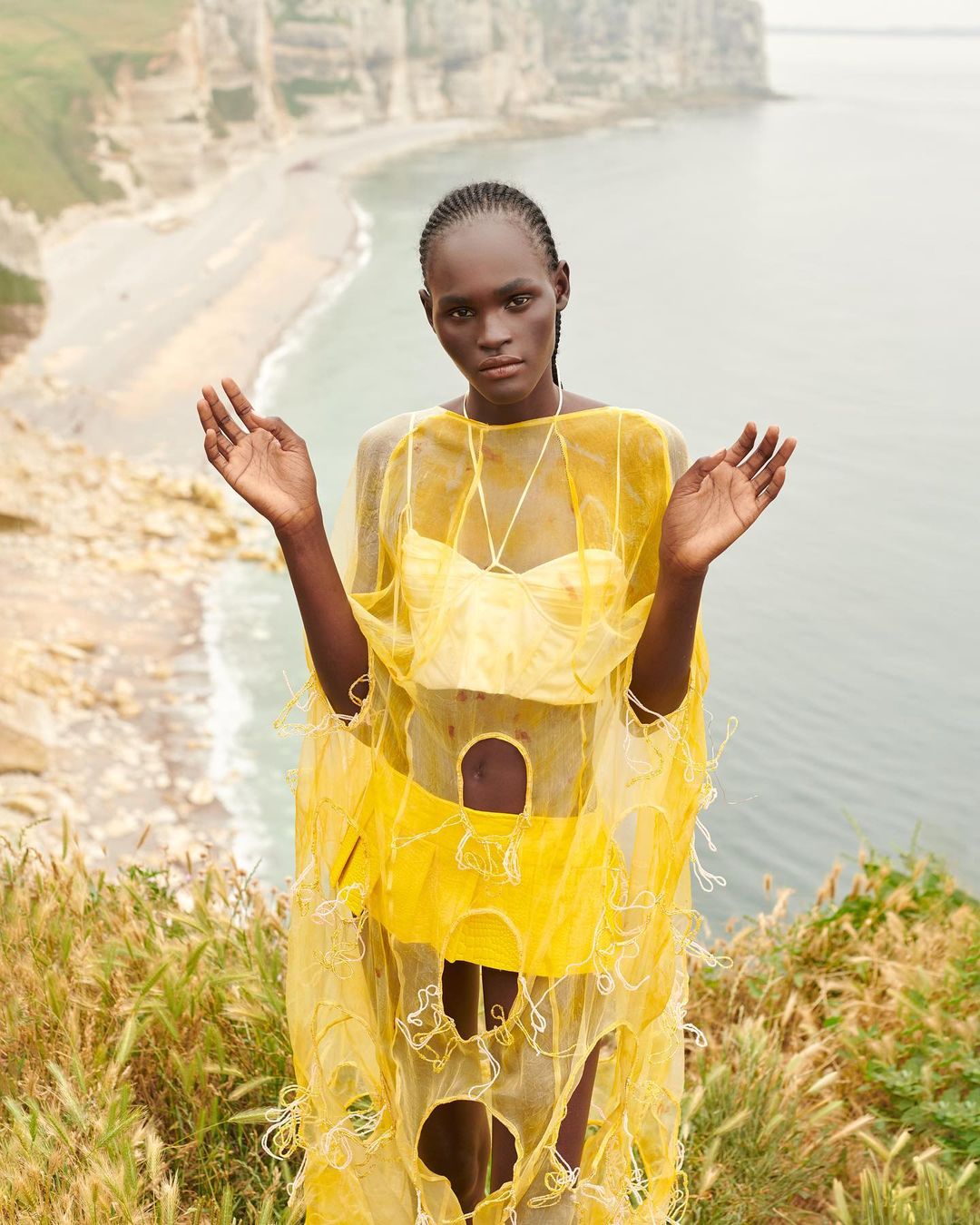 A striking editorial fashion portrait of an African woman in a yellow outfit, set against a backdrop of rocky cliffs and the sea.