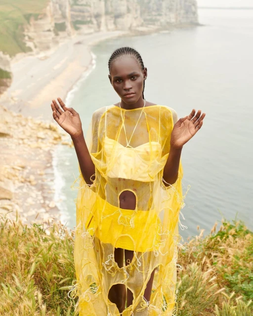 A striking editorial fashion portrait of an African woman in a yellow outfit, set against a backdrop of rocky cliffs and the sea.