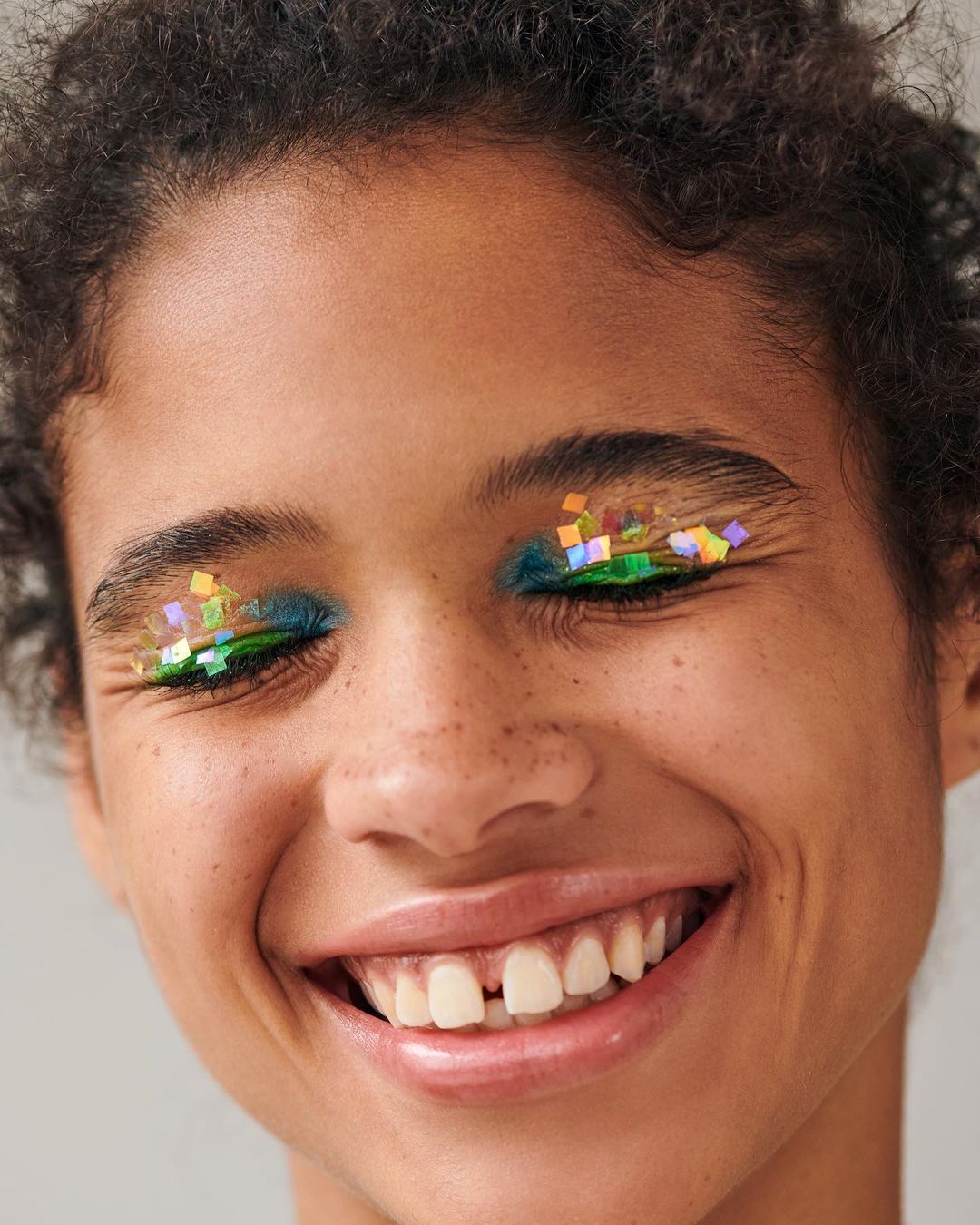 Close-up portrait of an African-American woman smiling with glitter eye makeup and dark curly hair against a neutral grey background.