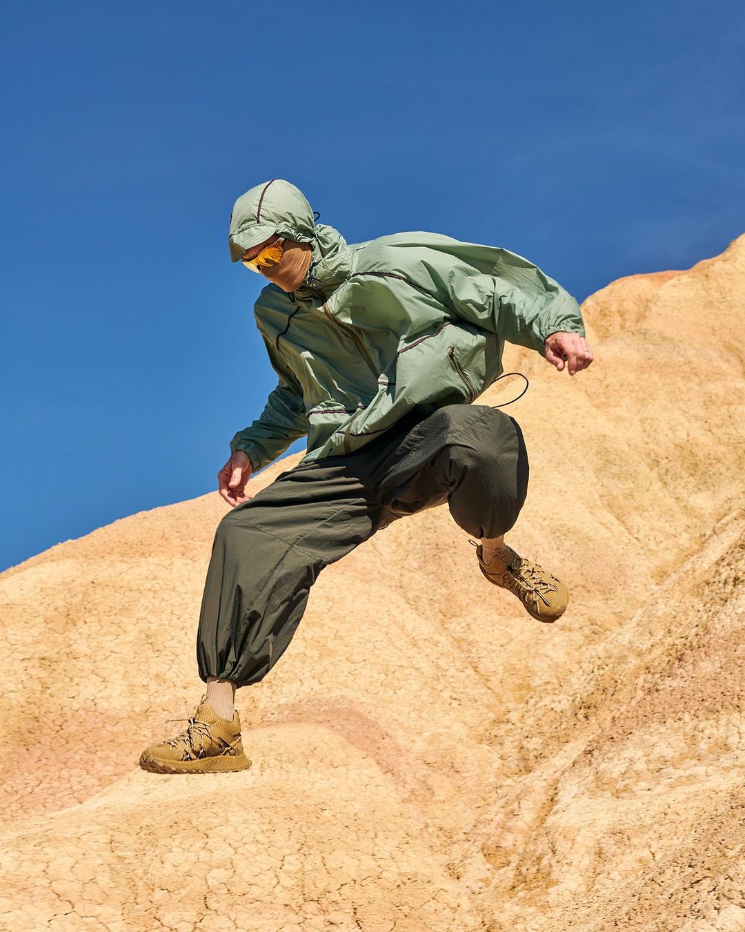 A person in an olive hooded jacket, dark baggy pants, and tan hiking boots jumps off a tan desert dune under a bright blue sky.