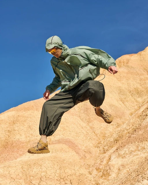 A person in an olive hooded jacket, dark baggy pants, and tan hiking boots jumps off a tan desert dune under a bright blue sky.