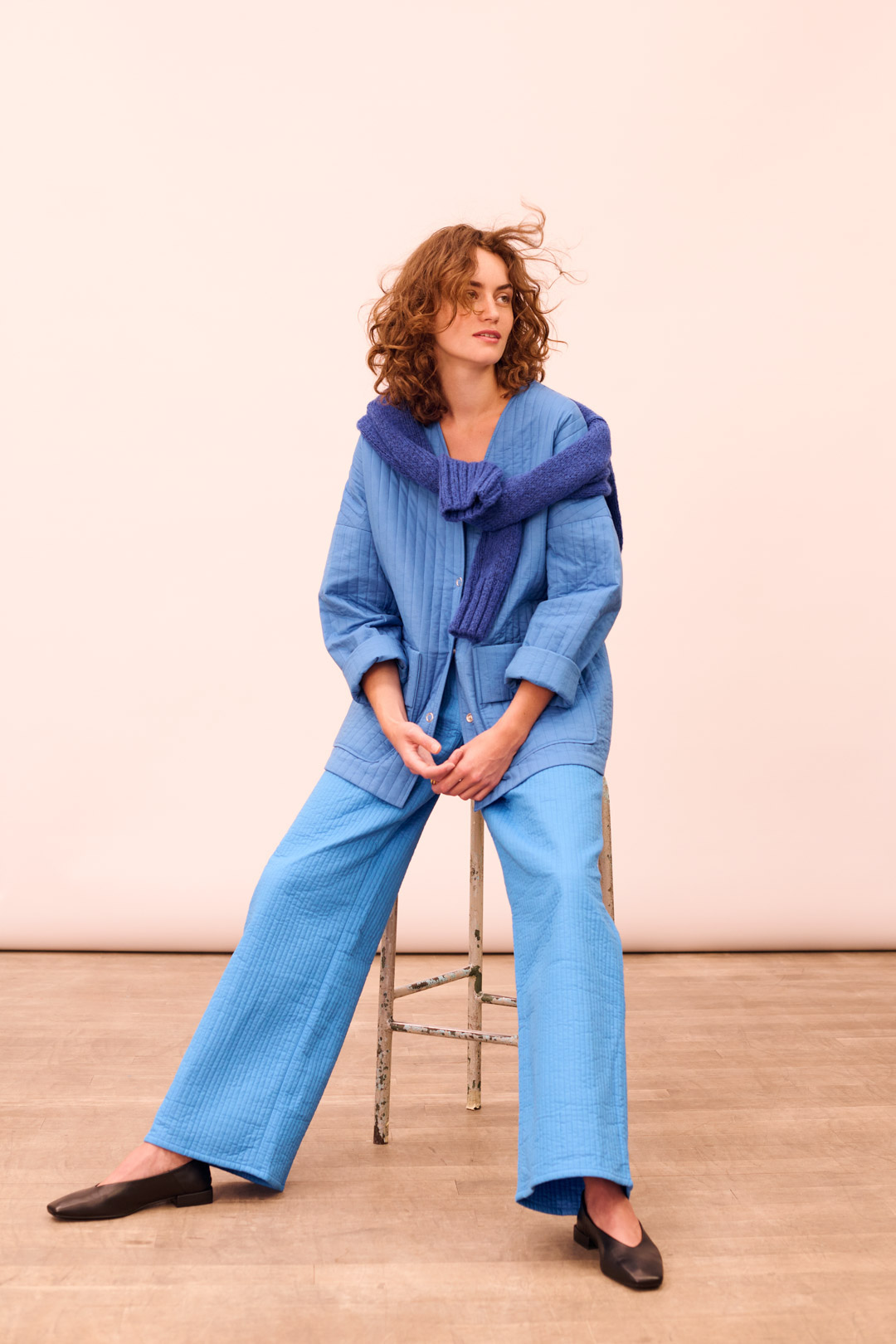 A stylish woman poses while seated on a stool, wearing a blue quilted suit and a blue sweater tied over her shoulders in a studio setting.