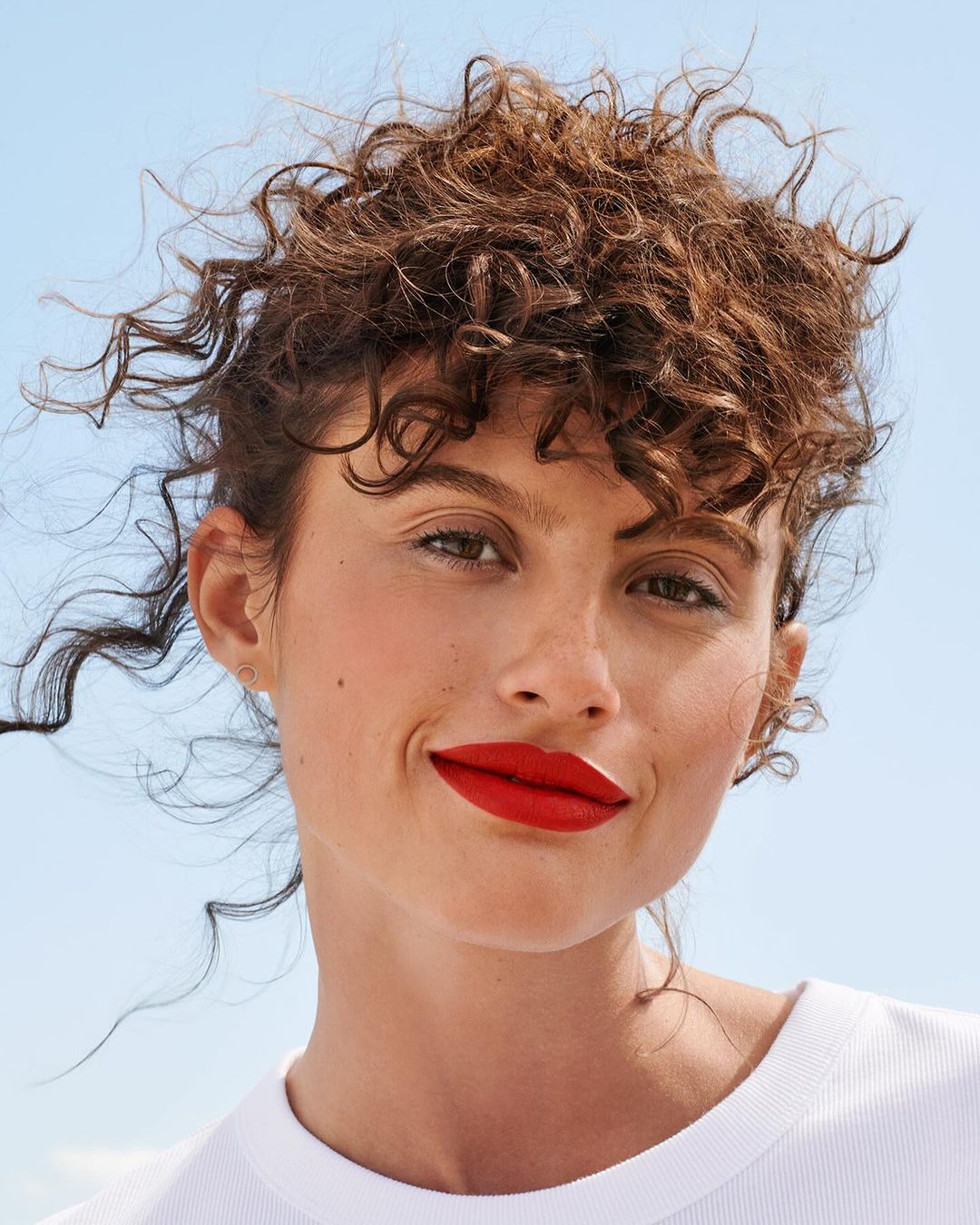 Close-up portrait of a woman with curly brown hair, red lipstick, and a white t-shirt against a clear blue sky