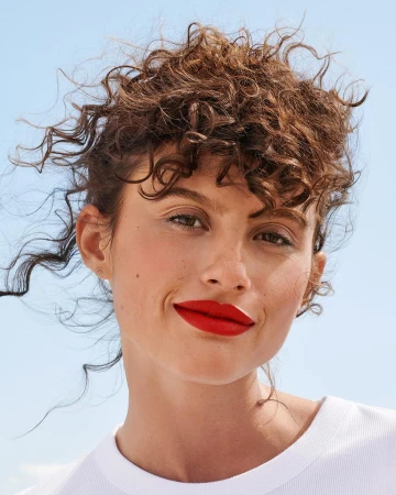 Close-up portrait of a woman with curly brown hair, red lipstick, and a white t-shirt against a clear blue sky