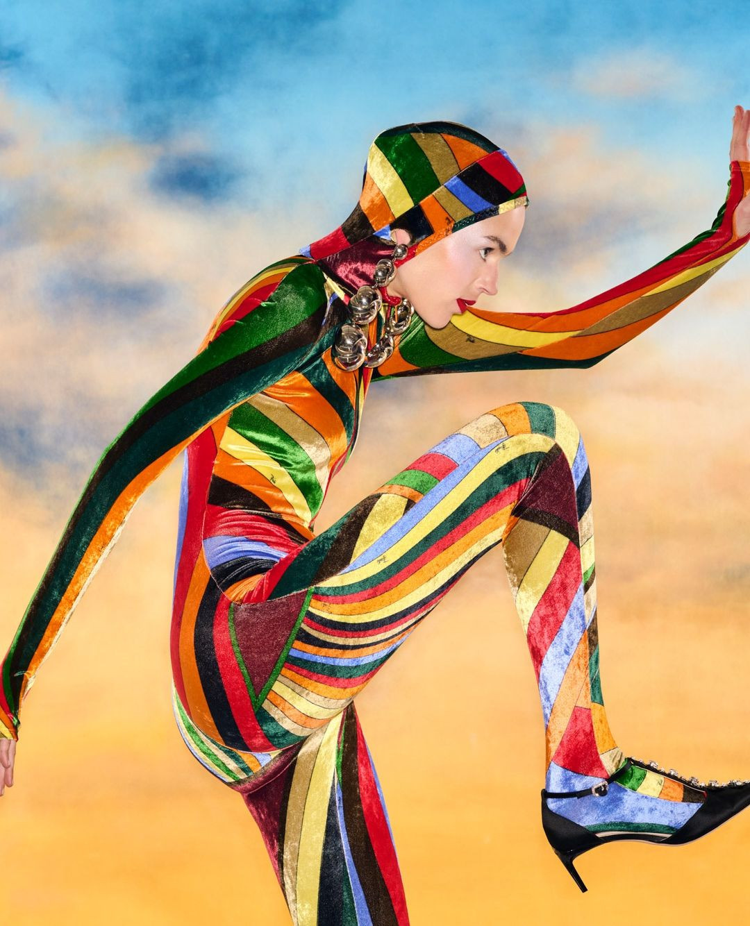 Fashion shot of a model in a multicolor patterned catsuit and hood posing in a dynamic stance against a cloudy sky background.