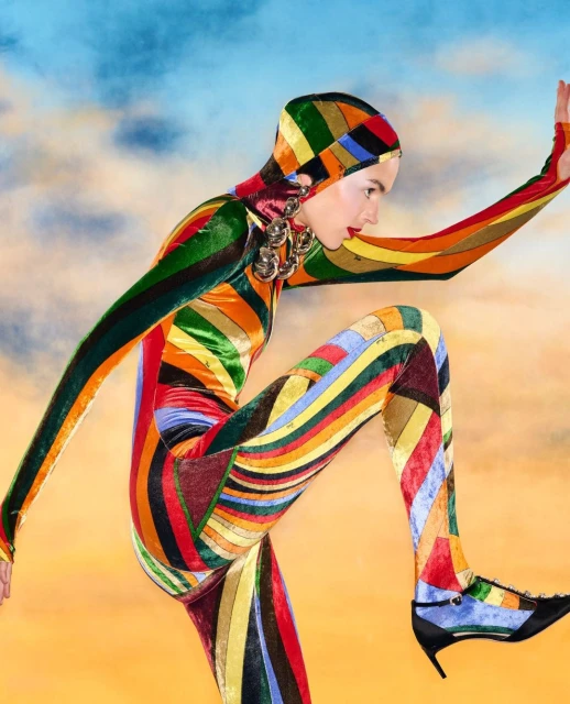 Fashion shot of a model in a multicolor patterned catsuit and hood posing in a dynamic stance against a cloudy sky background.