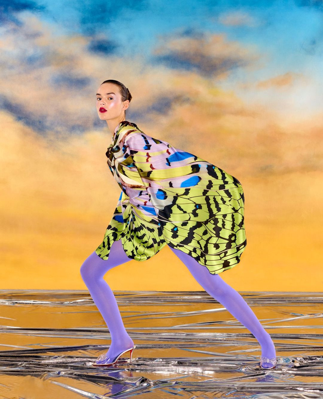A woman with slicked-back hair poses in a butterfly wing-patterned dress and purple tights against a surreal, dreamlike studio backdrop.