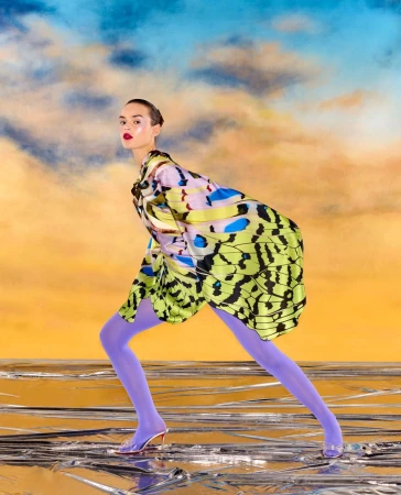 A woman with slicked-back hair poses in a butterfly wing-patterned dress and purple tights against a surreal, dreamlike studio backdrop.