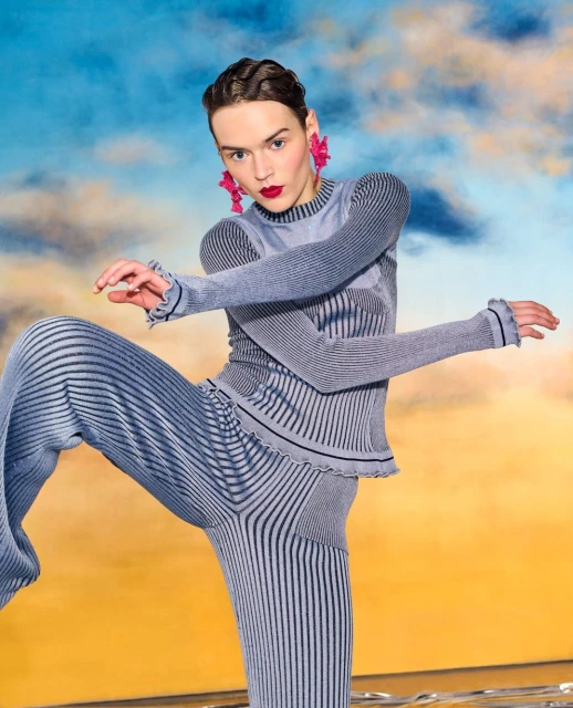 Model strikes a pose in blue ribbed knitwear against a dynamic sky backdrop, showcasing avant-garde fashion with bold red lipstick and earrings.