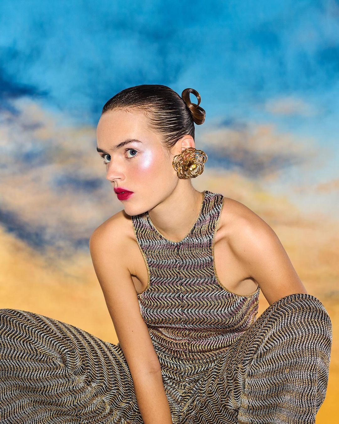 A woman poses in gold patterned sleeveless top and pants against a blue and orange cloudy background, wearing oversized gold earrings.