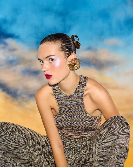 A woman poses in gold patterned sleeveless top and pants against a blue and orange cloudy background, wearing oversized gold earrings.