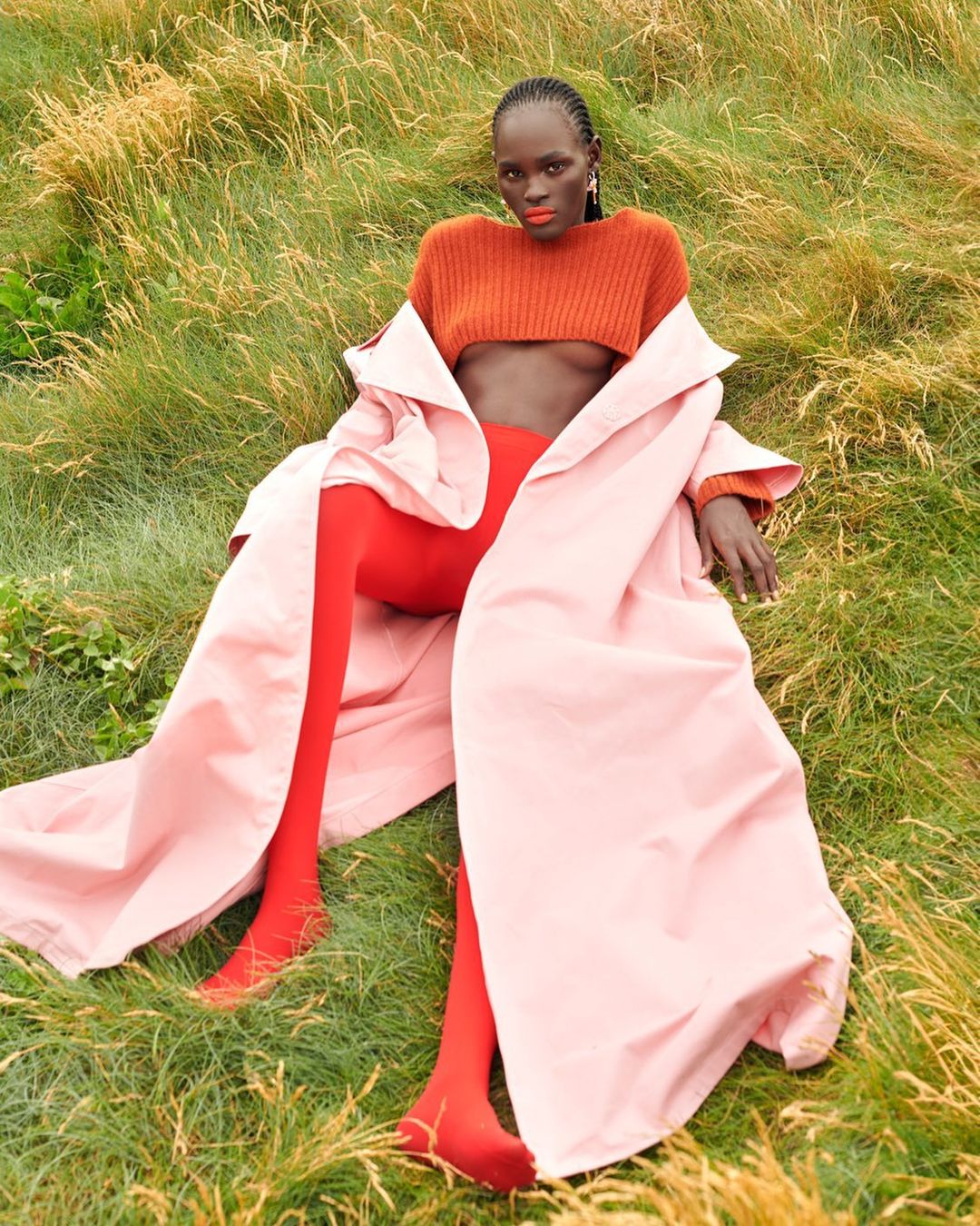 Woman in orange crop top, pink coat, and red tights lying in tall grass. Fashion editorial outdoor shot with green and golden tones.