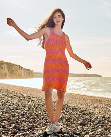 Woman in a pink and orange striped knit dress standing on a pebble beach by the ocean during sunset