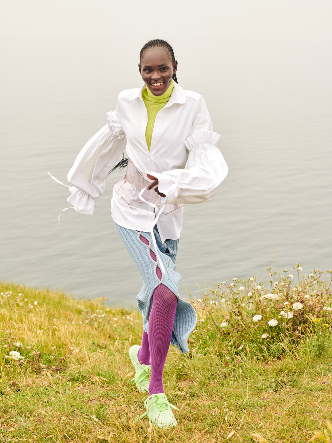 A cheerful African woman in a stylish white shirt and skirt walks on a grassy hill with the ocean and sky in the background.
