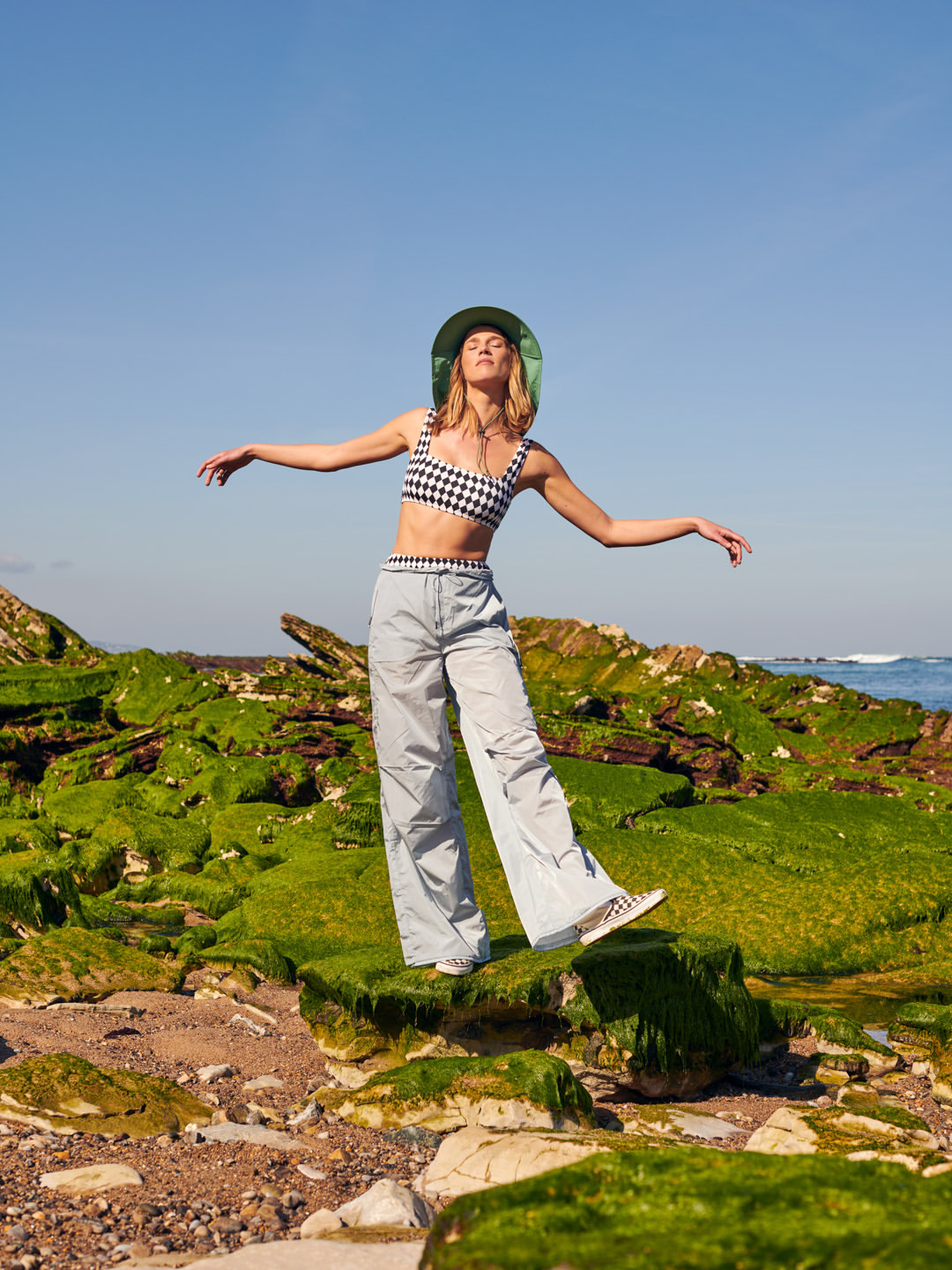A woman in a hat, patterned top, and wide-leg pants balances on moss-covered rocks near the ocean under a clear blue sky on a sunny day.