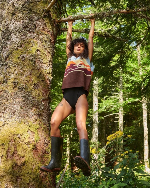 Woman in top and black briefs hangs from tree branch outdoors in sunlight, wearing rubber boots in a forest setting.