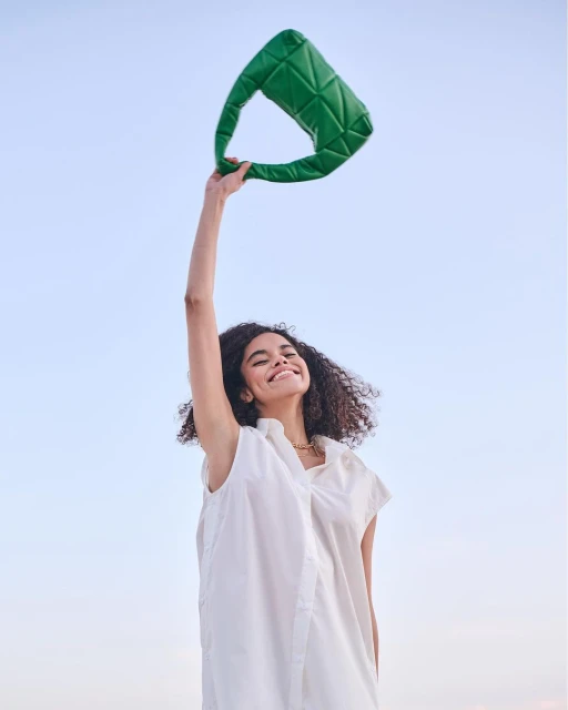 Smiling woman with curly hair in a white dress raises a green, quilted handbag against a light blue sky, capturing a joyous outdoor moment.