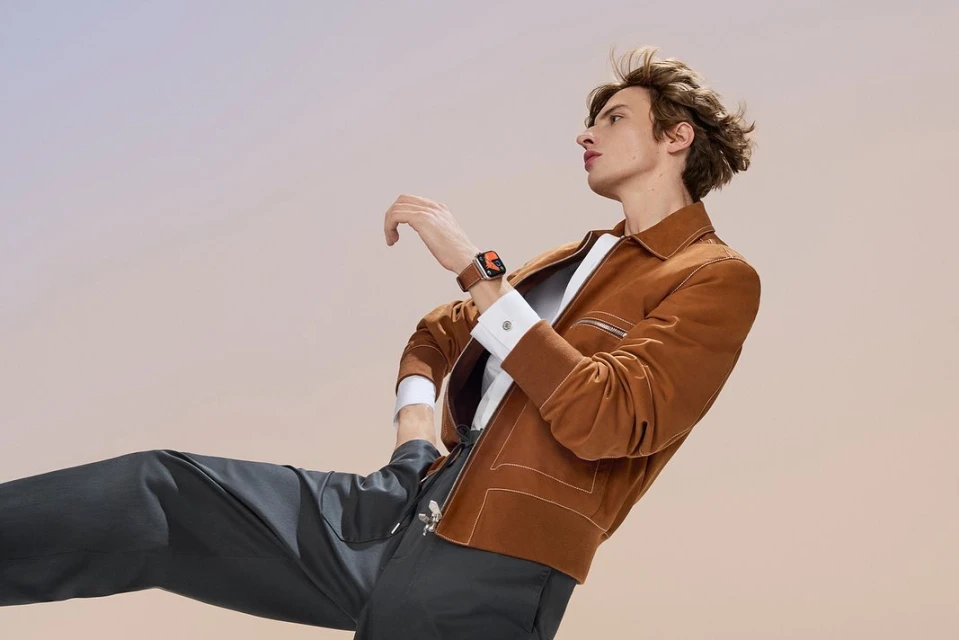 An adult male wearing a brown jacket and black pants poses at an angle in front of a light-colored backdrop, showcasing a wrist watch.