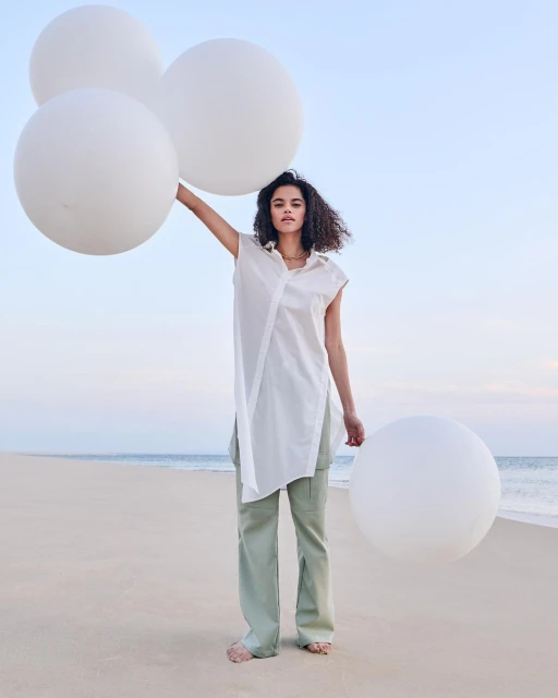 Woman in a white shirt and green pants stands barefoot on a sandy beach, holding three large white balloons against a blue sky.