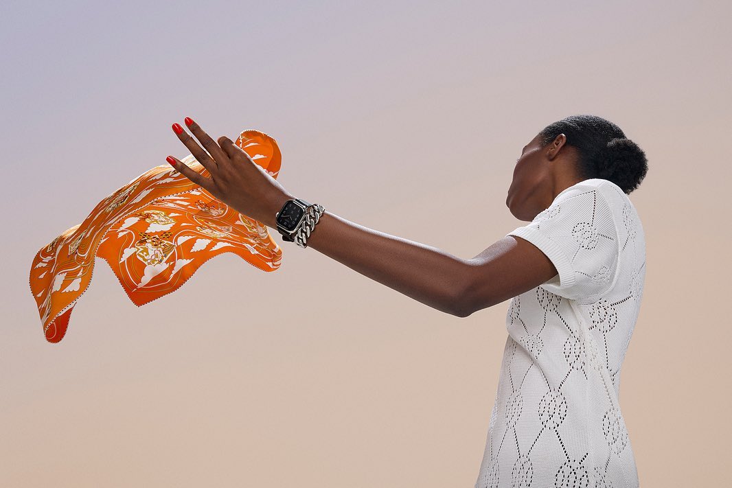 Woman in a white textured dress holds up an orange patterned scarf, showcasing a smartwatch with a silver chain band against a soft gradient backdrop.