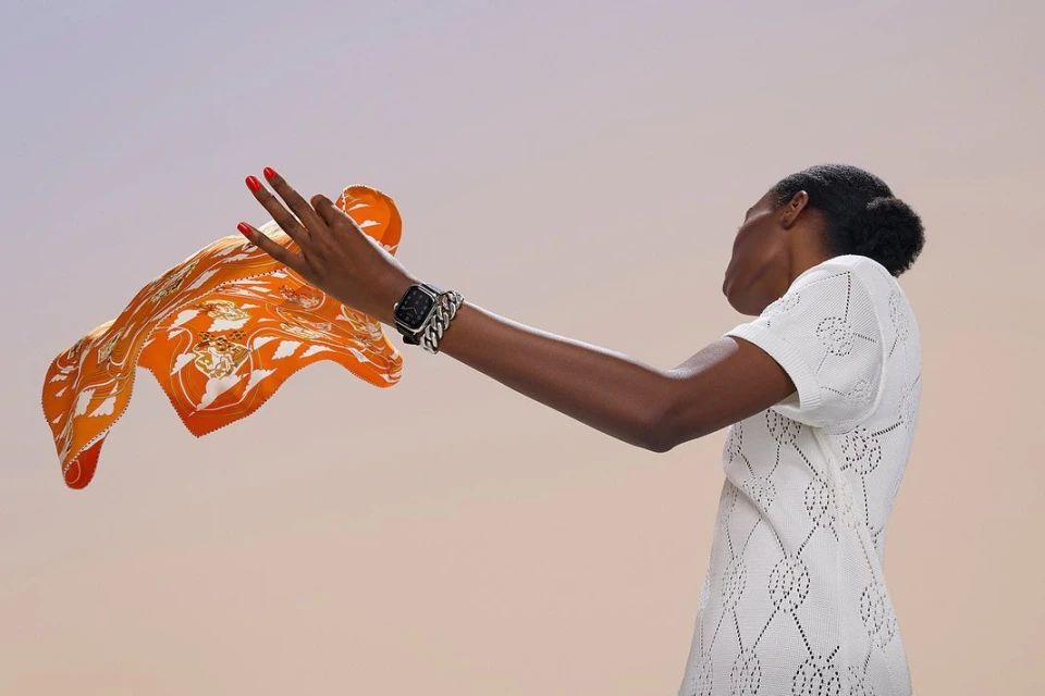 Woman in a white textured dress holds up an orange patterned scarf, showcasing a smartwatch with a silver chain band against a soft gradient backdrop.