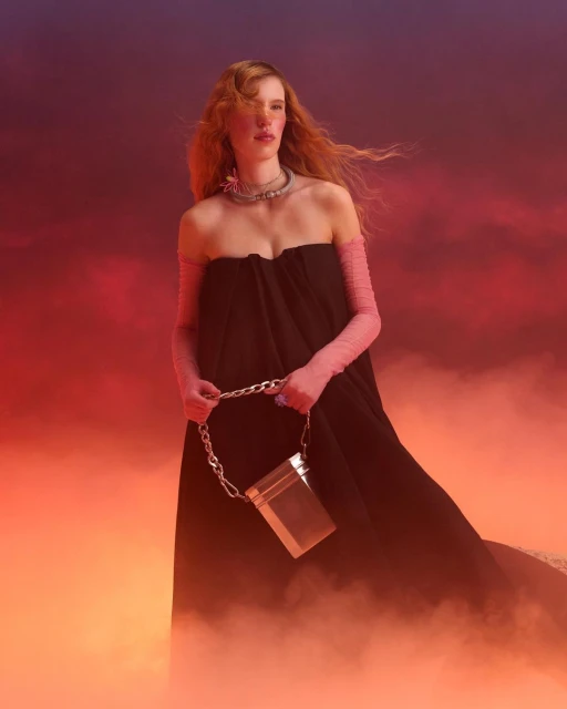 Elegant woman in black dress holds silver bag against a hazy red and orange background. Fashion photography, studio lighting, portrait shot.
