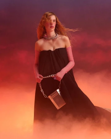 Model with red hair wearing a black dress and pink sleeves holding a metallic bag against a fiery sunset sky.