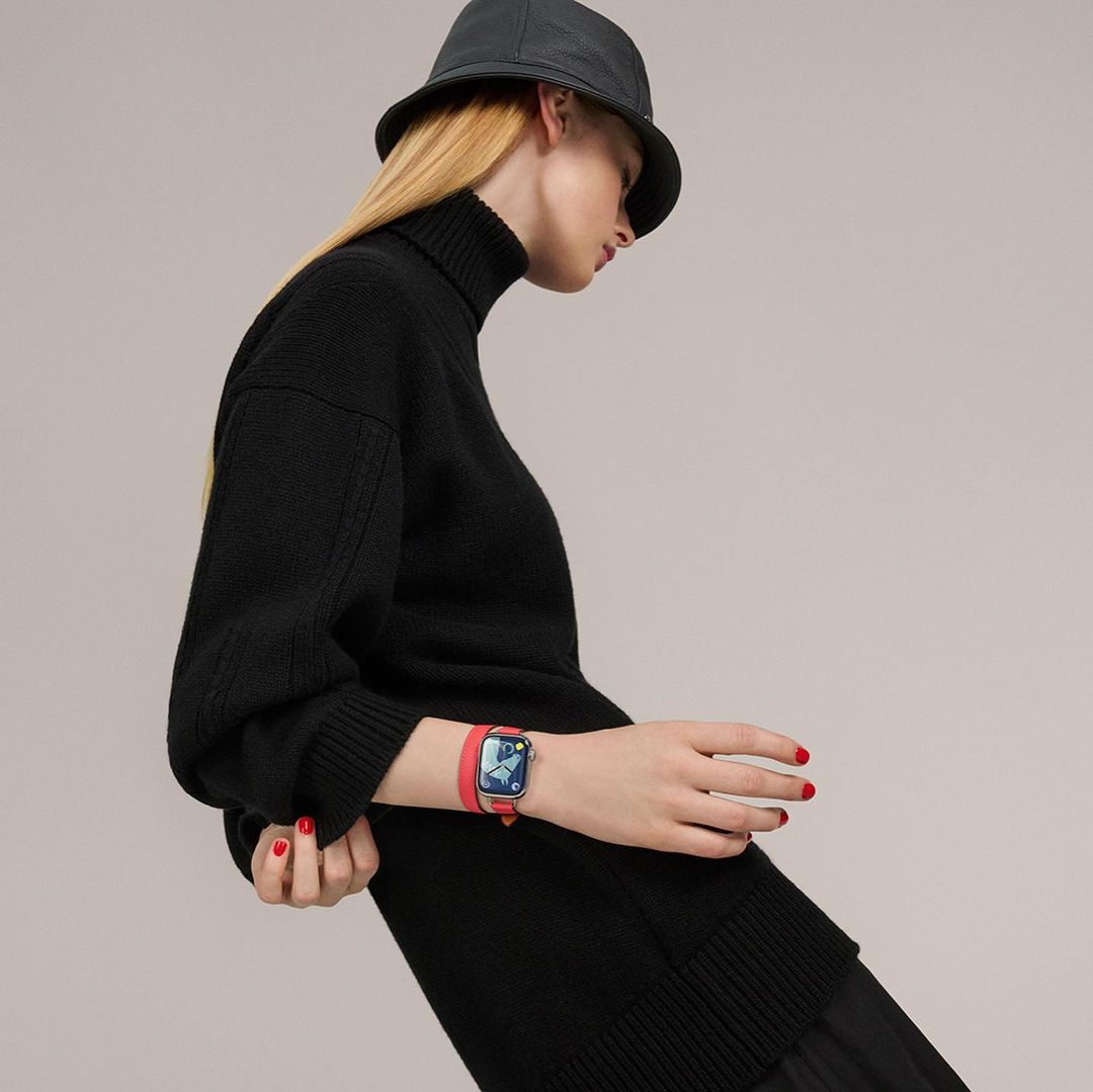 A blonde woman wearing a black turtleneck sweater, bucket hat, and a red smart watch, stands against a neutral background in a studio shot.