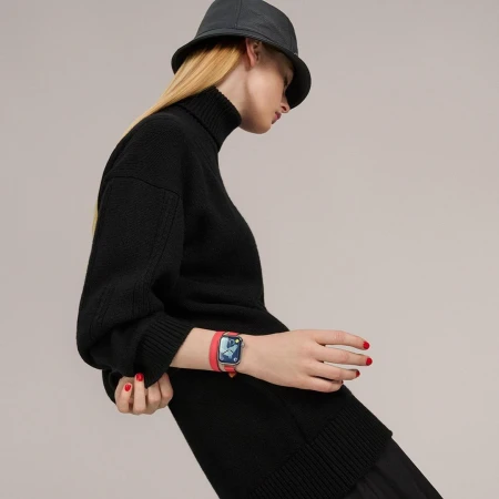 A blonde woman wearing a black turtleneck sweater, bucket hat, and a red smart watch, stands against a neutral background in a studio shot.