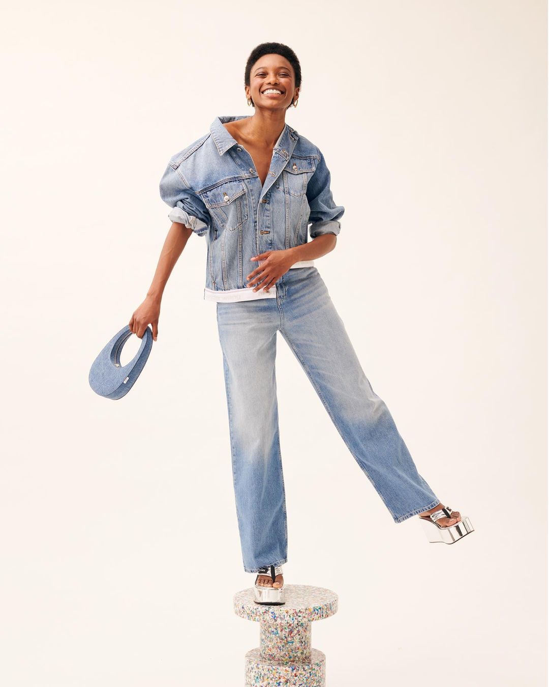 Smiling woman in oversized denim jacket and jeans standing on a terrazzo pillar holding a small blue handbag