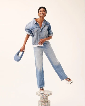 Smiling woman in oversized denim jacket and jeans standing on a terrazzo pillar holding a small blue handbag