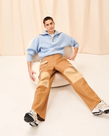 An adult male wearing a blue quarter-zip sweater and tan carpenter pants poses indoors against a cream-colored backdrop in a studio shot.