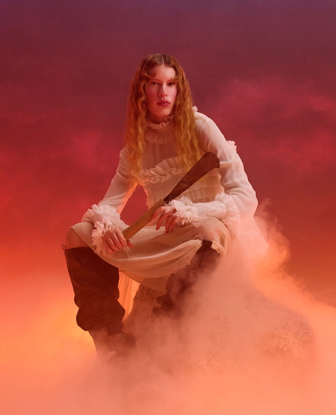 Woman in a white dress holds a machete, seated in a hazy environment with an orange and purple sky background, creating an ethereal atmosphere.