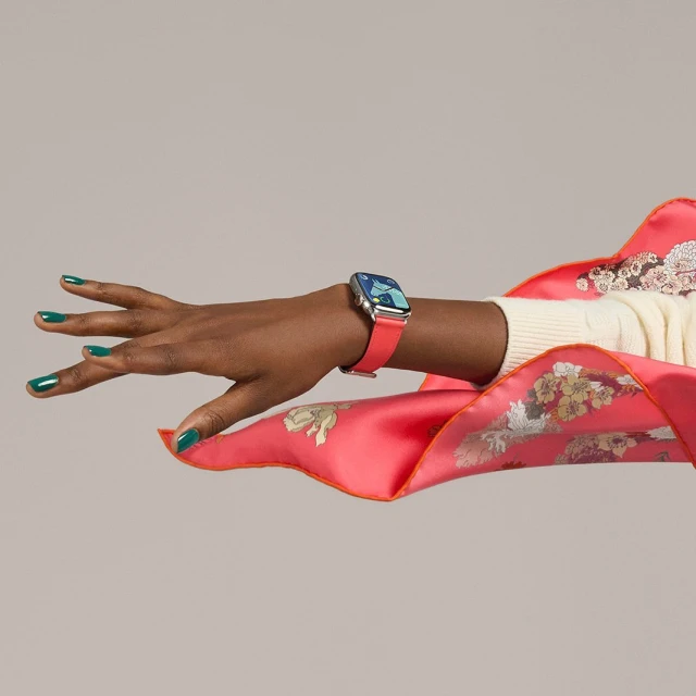 Close-up of a dark-skinned hand wearing a smart watch with a pink band, accessorized with a patterned silk scarf and dark green nail polish.