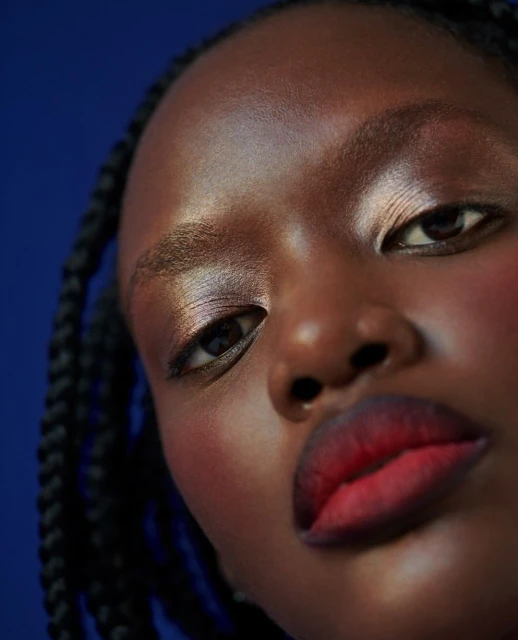 Close-up portrait of a Black woman with gradient red lips, iridescent eyeshadow, and braided hair against a deep blue background.